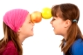 Laughing small girls facing each other, holding two apples together with forehead.�