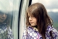 Sad beautiful little girl leaning against a window standing thinking with downcast eyes reflected in the glass